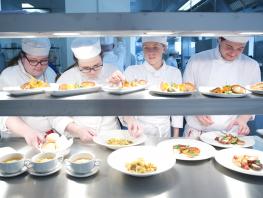 Culinary students standing in row behind counter preparing plates for serving. 
