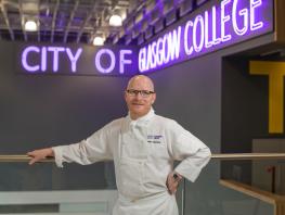 Gary Maclean standing against railing with neon sign in background saying City of Glasgow College 