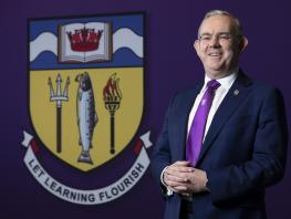 Principal Paul Little standing next to College crest which says Let Learning Flourish 