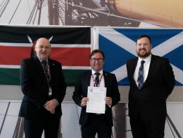 Three members of staff standing in front of a Kenyan and Scottish flag.  Middle person is holding a piece of paper. 