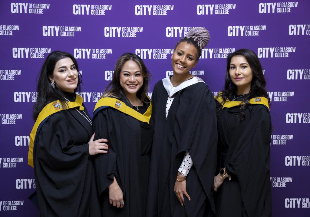 Four students standing in front of a college stand in academic robes.