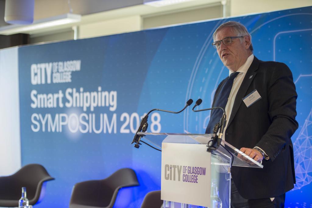 Captain John Lloyd standing at the lectern with a blue Smart Shipping Symposium background.