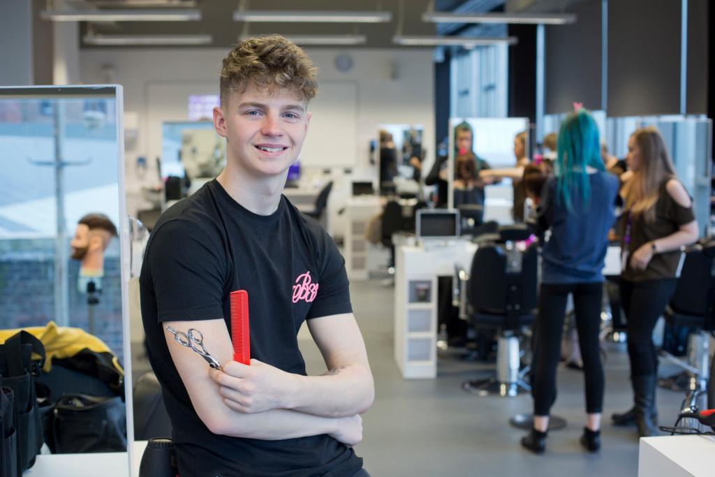 Hairdressing student sitting on the edge of a desk holding a scissors and a red comb.  Other students are in the background working on clients hair.