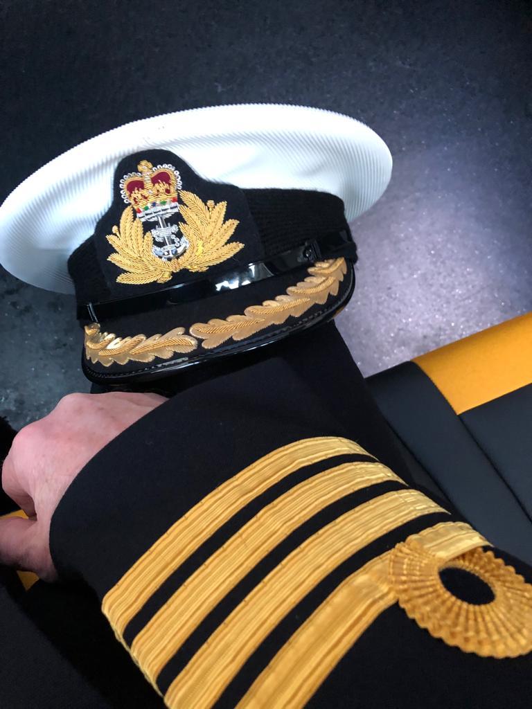 A white naval hat with black and gold design and the arm of a black jacket with four gold stripes.