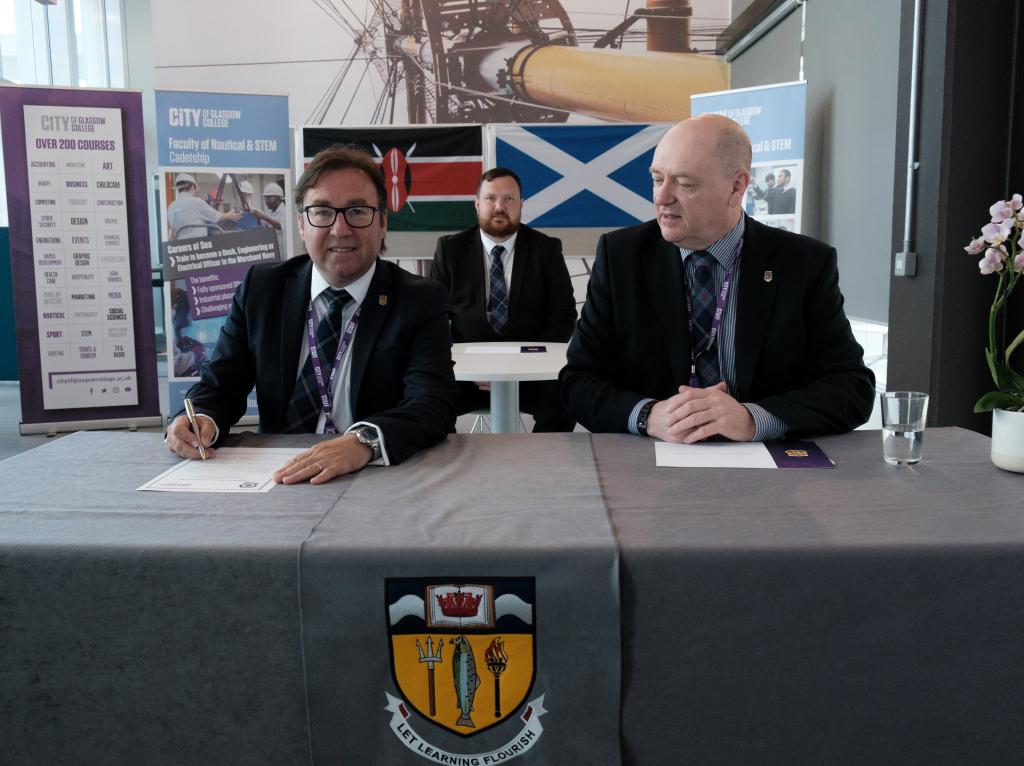 Two people sitting at a desk signing the memorandum with one person sitting at a desk behind them.