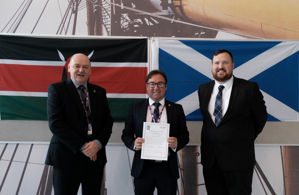 Three members of staff standing in front of a Kenyan and Scottish flag.  Middle person is holding a piece of paper.