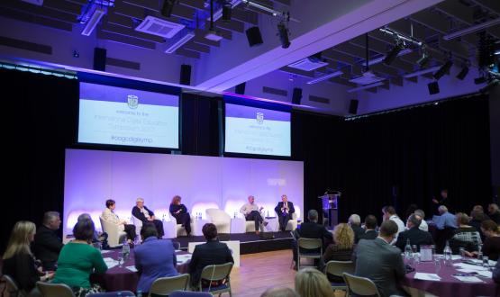 Lecture theatre set up for a conference with roundtables and stages with five people sitting on it.  In the background against the wall are two televisions.