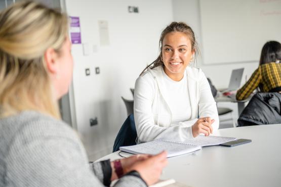 Two students sitting at a desk in a classroom chatting.