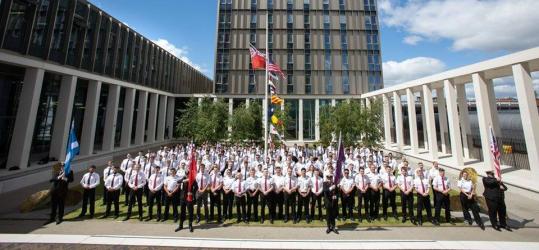 A big group of cadet sin uniform lined up in a terrace area at Riverside campus with the accommodation block in the background and flags flying.