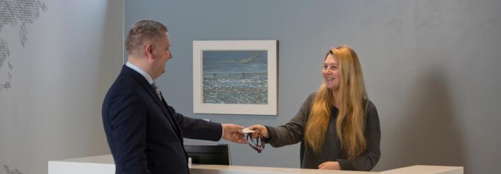 Two people standing at a reception desk.  The person behind the desk is handing the person in front of the desk a badge.