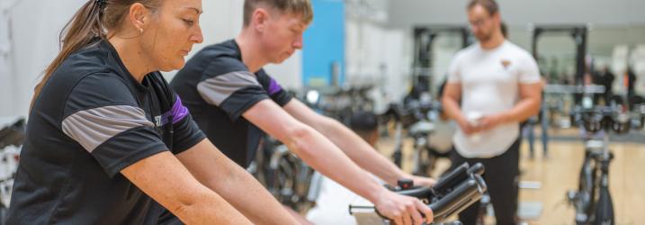Two students on spin bikes in a gym with another student standing in the background