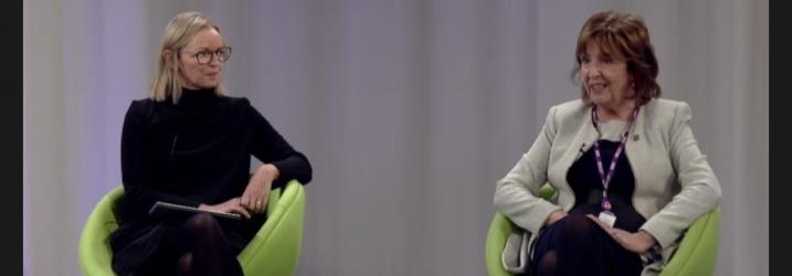 Two panelists sitting in green chairs on a stage with a white background.