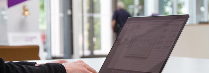 A laptop sitting on a table with someone typing on it.  You can only see their hands.