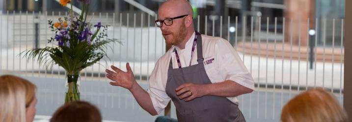 Gary Maclean in chefs whites and apron standing in front of a window talking to an audience