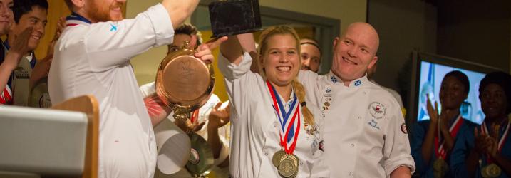 Three chefs standing wearing medals and holding a cup up