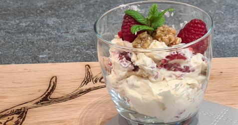 A glass sitting on a wooden board with Cranachan in it.  Cream and oats with on the bottom with raspberries and a sprig of fresh mint on the top.