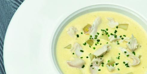 A close up of a white bowl with cullen skink in it.  There is a yellow coloured soup with flaked bits of fish sprinkled with green herbs.