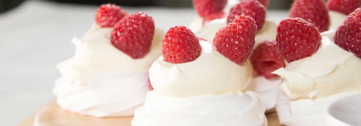MEringue nests sitting on a round wooden board topped with cream and raspberries.