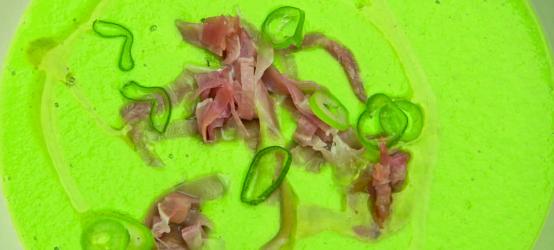 A white bowl with green pea and ham soup topped with bits of ham and spring onions.
